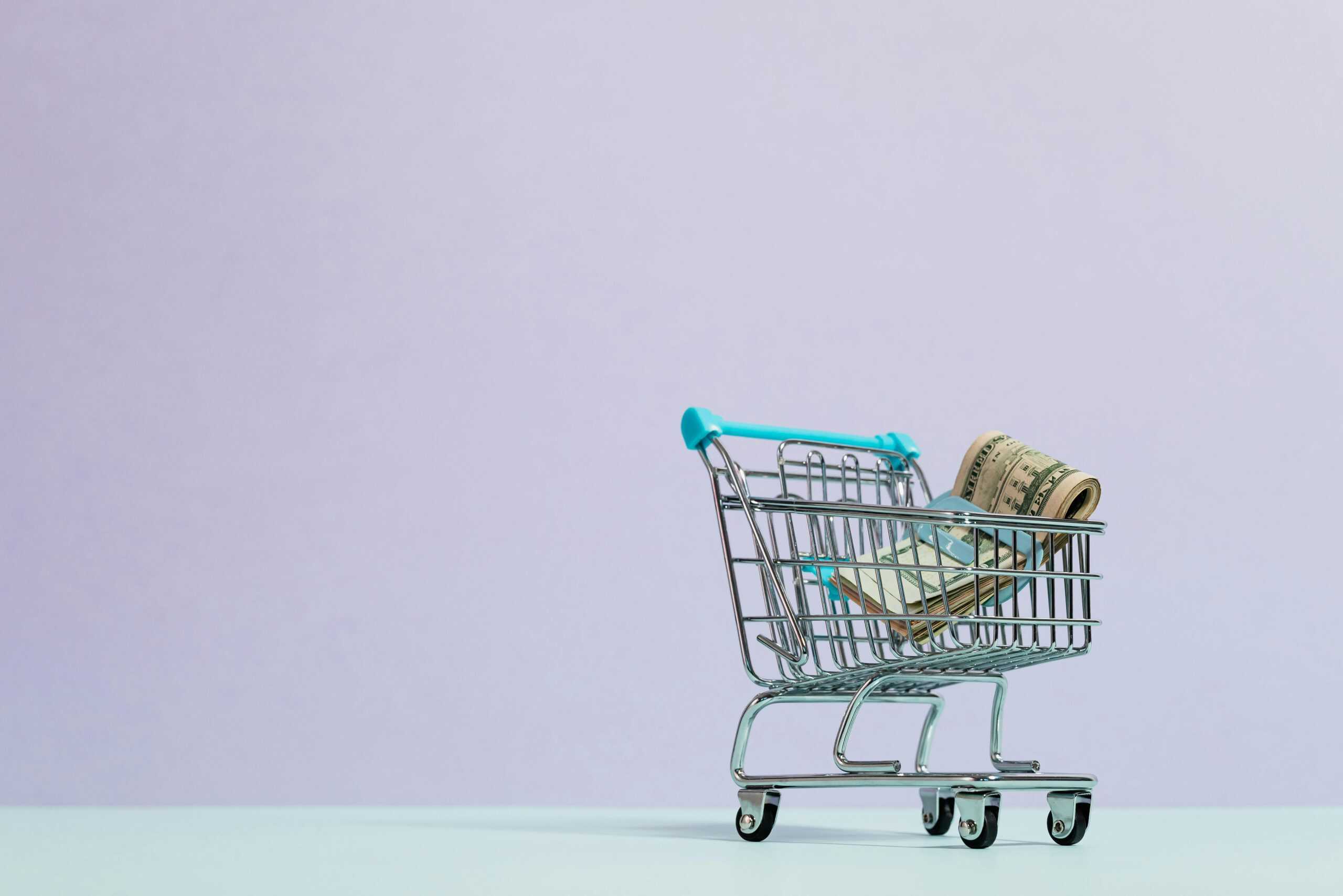 Mini shopping cart filled with rolled-up U.S. dollar bills on a pastel background, representing spending and saving habits fueled by the no-spend challenge