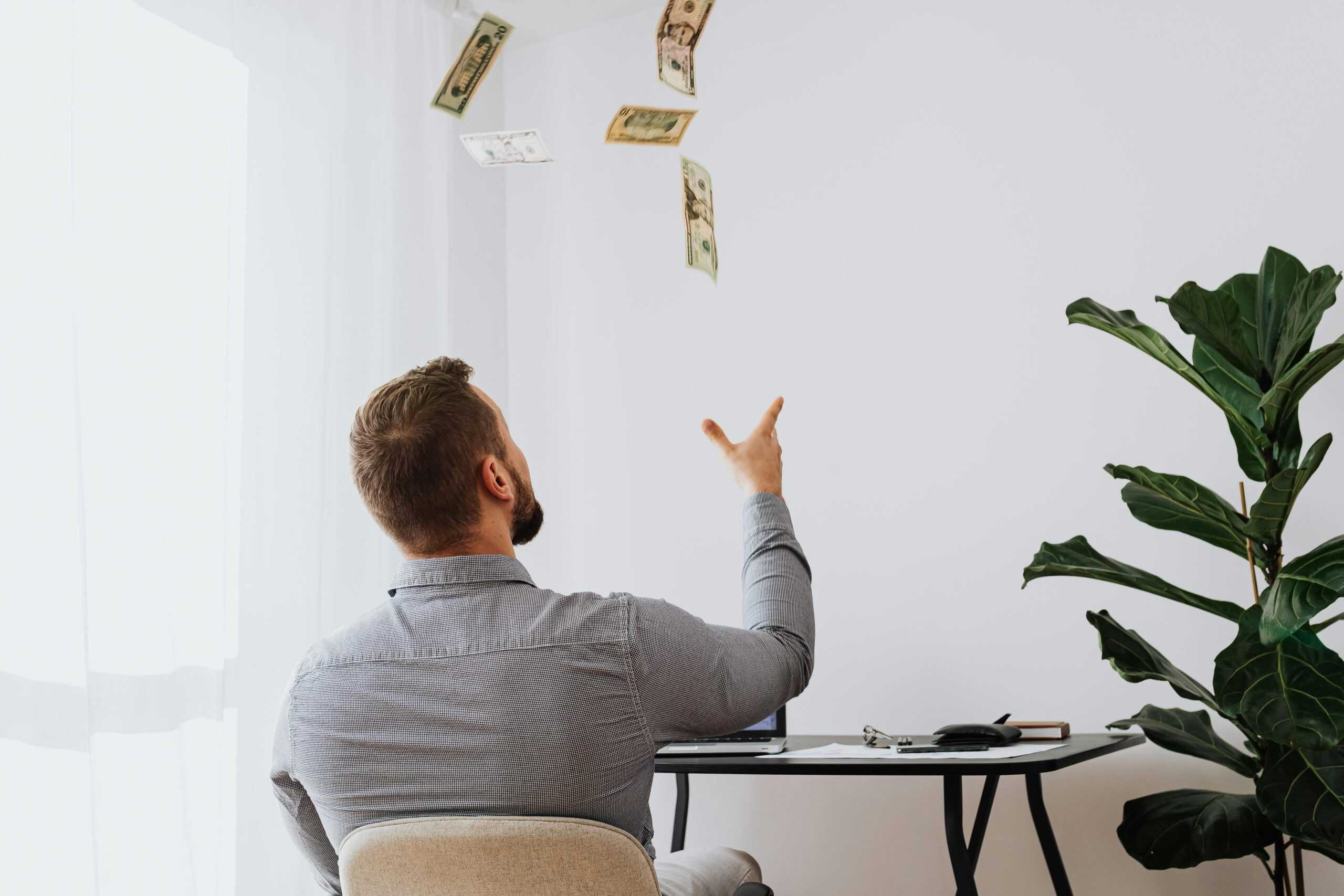 Man in a gray shirt tossing money into the air while sitting at a desk, symbolizing financial freedom and a money mindset shift.