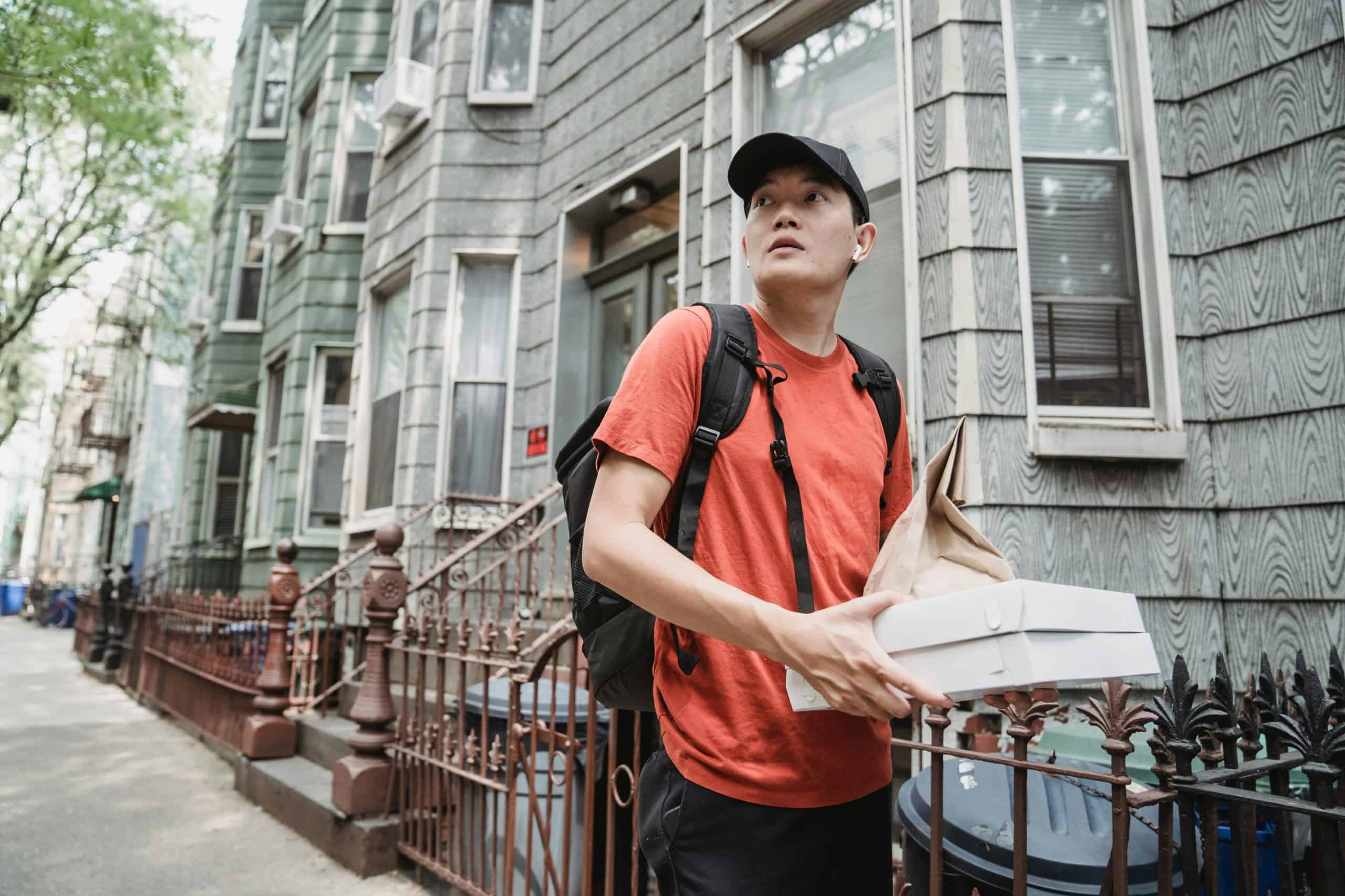 Delivery worker wearing a red shirt and backpack holding food packages in front of apartment buildings — representing gig economy life and budgeting for irregular income.